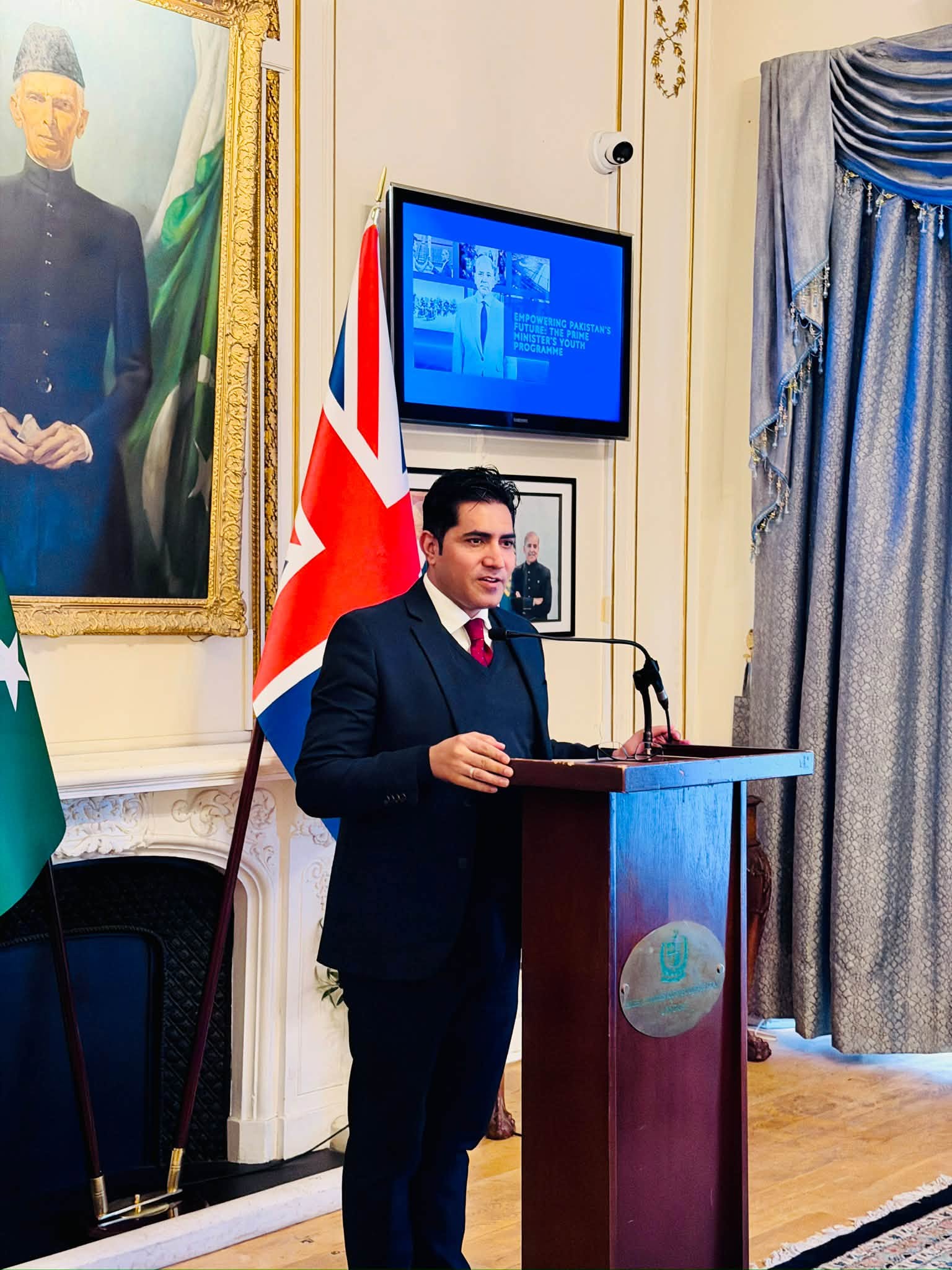 Speaker at the podium beside a screen titled Empowering Pakistan's Future: The Prime Minister's Youth Programme at the Pakistan High Commission in London