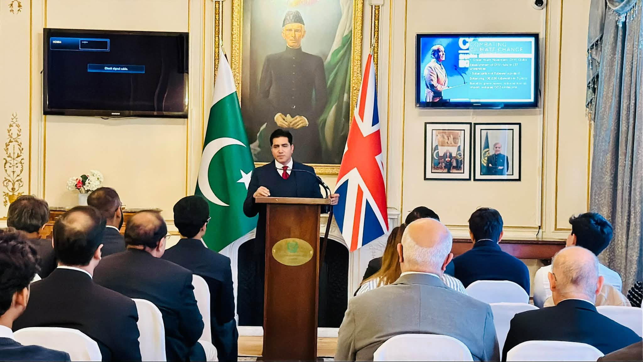 Speaker at a podium presenting to an audience at the Youth Engagement Session at the Pakistan High Commission in London, with a slide on combating climate change